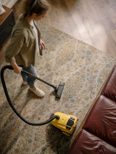 A person operating a yellow upright vacuum cleaner with a black top and hose, performing surface cleaning on a patterned carpet in a residential living room. The vacuum's dust compartment is open, and