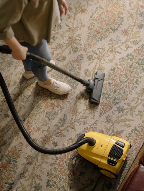 A person vacuuming a patterned, beige and pastel-colored carpet in a room with wooden flooring visible at the edge. The individual, wearing beige shoes and blue jeans, is using a yellow and black vacuum cleaner with a long metal wand to apply surface cleaning and deep cleaning for sanitisation. The scene is well-lit, highlighting the clean condition of the carpet, with the vacuum cleaner's hose connected to the main unit on the floor, situated in a domestic setting. Carpet Cleaning SW6 specializes in professional cleaning services, ensuring hygiene and freshness in residential spaces along Fulham Broadway and New Kings Road.