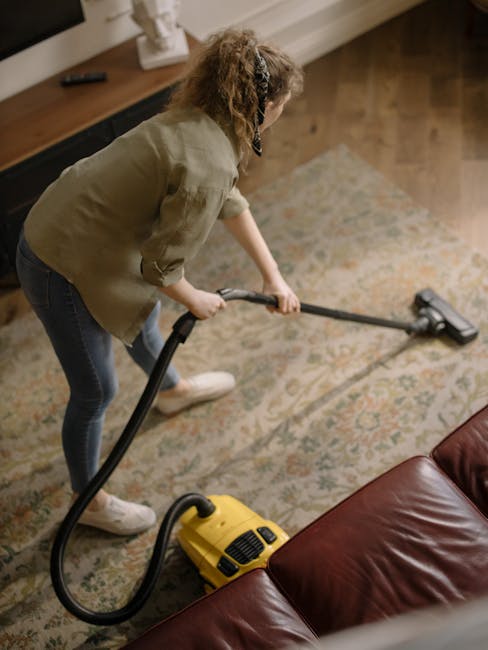 A woman engaged in deep surface cleaning using a vacuum cleaner with a hose attachment on a patterned area rug in a living room. She is wearing casual clothing including a beige jacket and dark jeans, standing on a wooden floor adjacent to the rug. The room features natural lighting, with a wooden media console and a white decorative object on top nearby. The scene emphasizes thorough hygienic maintenance, demonstrating professional carpet cleaning services provided by Carpet Cleaning SW6, aligned with the focus on deep cleaning and sanitisation in Fulham Broadway and New Kings Road, SW6.