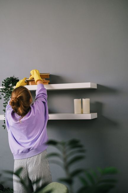 A person wearing yellow rubber gloves and a purple sweater is reaching up to adjust or clean a wooden object on a white floating shelf mounted on a light grey wall in a domestic setting. The shelves h