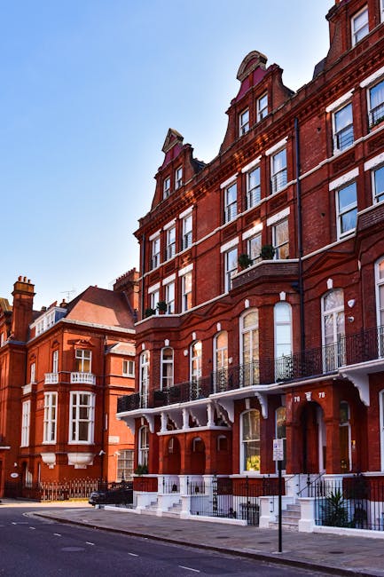 A row of Victorian-style terraced houses with ornate architectural details, painted in various pastel shades including blue, beige, and green. The buildings feature large, double-hung sash windows wit
